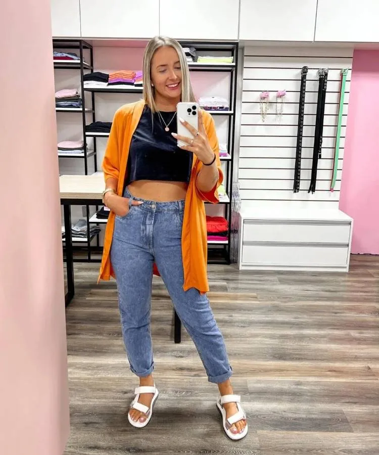 Uma mulher branca e loira posa em frente a um espelho, ela usa calça jeans, blusa peta e um kimono laranja, nos pés uma papete branca