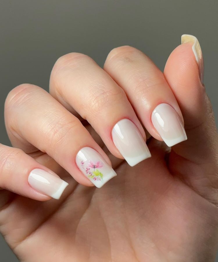 A imagem mostra uma mão com unhas bem cuidadas em um estilo de francesinha, com esmalte branco nas pontas. Uma das unhas possui uma decoração floral delicada em tons de rosa e verde, adicionando um toque de cor. Essa é uma unha decorada simples, que combina elegância e sutileza.