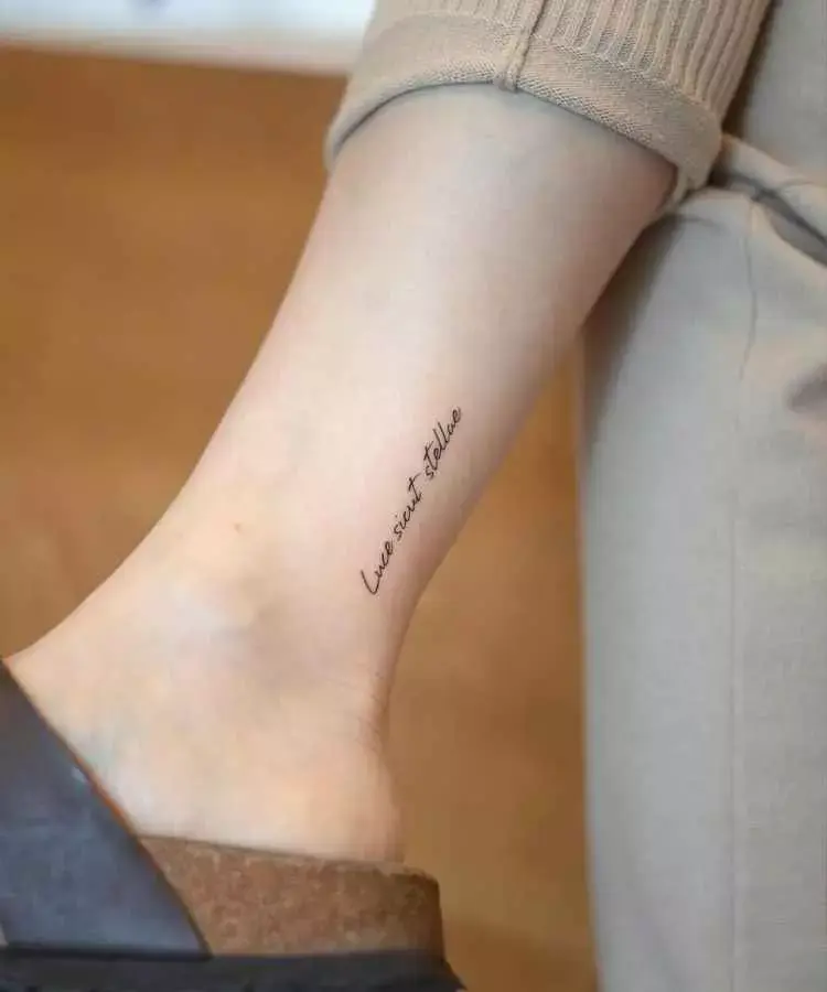 A imagem mostra uma tatuagem delicada no tornozelo com a frase em latim "luce siant stellae", escrita em uma fonte fina e cursiva. O fundo é desfocado, e a pessoa veste roupas em tons neutros, ressaltando a simplicidade elegante do design.