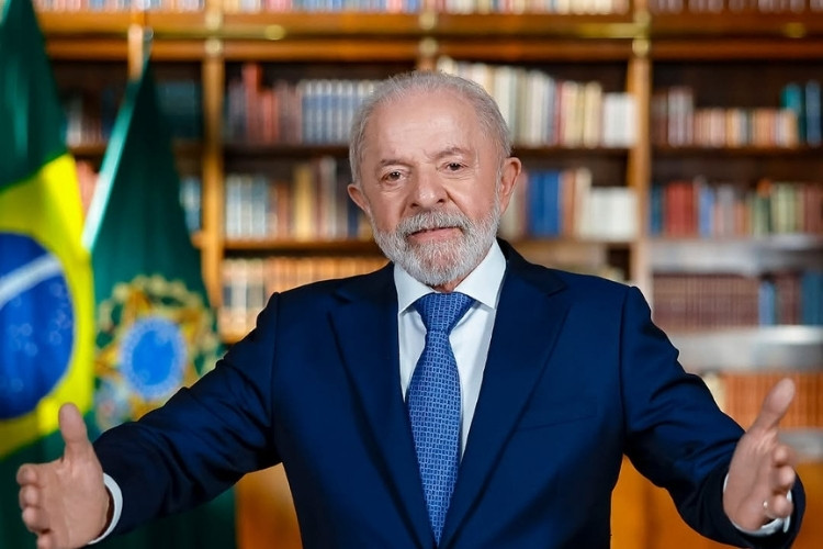 Presidente Luiz Inácio Lula da Silva, com barba e cabelos grisalhos, em um terno azul-marinho e gravata azul estampada. Ele está de pé, sorrindo, com os braços abertos em um gesto acolhedor, em frente a uma biblioteca.







