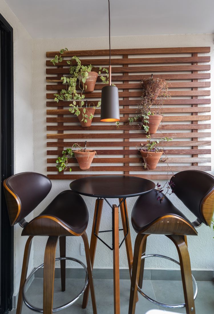A imagem mostra um ambiente de varanda com um painel de madeira ripado na parede, onde estão pendurados vasos de barro com plantas. Em frente ao painel, há uma mesa alta redonda de madeira escura, acompanhada de três banquetas com assentos curvos, em tom marrom e detalhes em madeira. Acima da mesa, uma luminária pendente moderna, de cor escura com o interior alaranjado, adiciona um toque de sofisticação. O espaço é compacto e aconchegante, com uma combinação de elementos naturais e modernos, ideal para momentos de descontração em um ambiente externo.


