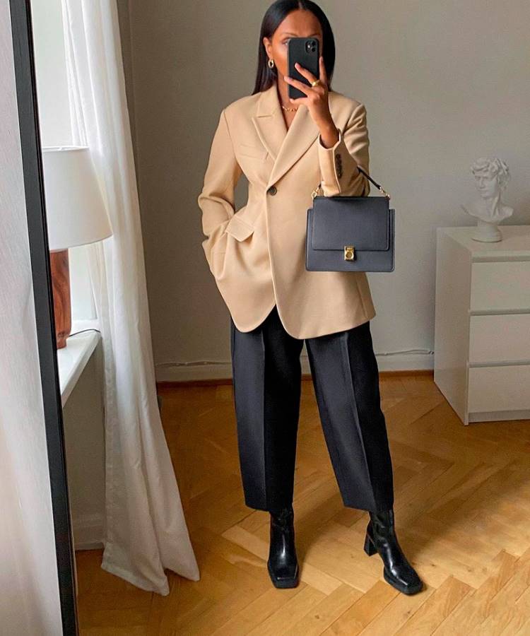 Mulher posa em um quarto minimalista vestindo blazer bege, calça preta reta e botas de couro pretas. Segura bolsa estruturada preta e celular. Cenário elegante com piso de madeira e móveis claros. Inspiração para looks de trabalho sofisticados.





