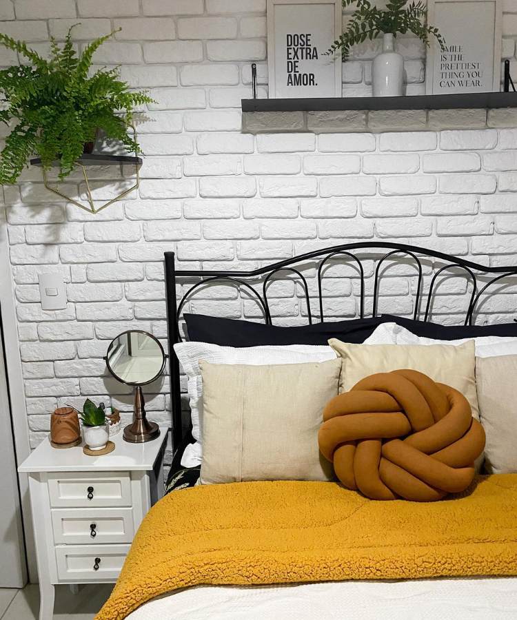 Quarto com parede de tijolinhos, cama de casal e planta samambaia