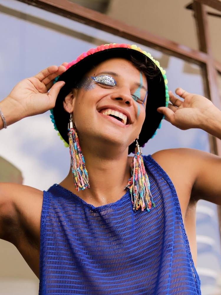 Homem com chapéu de pompons, brincos de paetês e regata azul transparente + make com glitter nos olhos