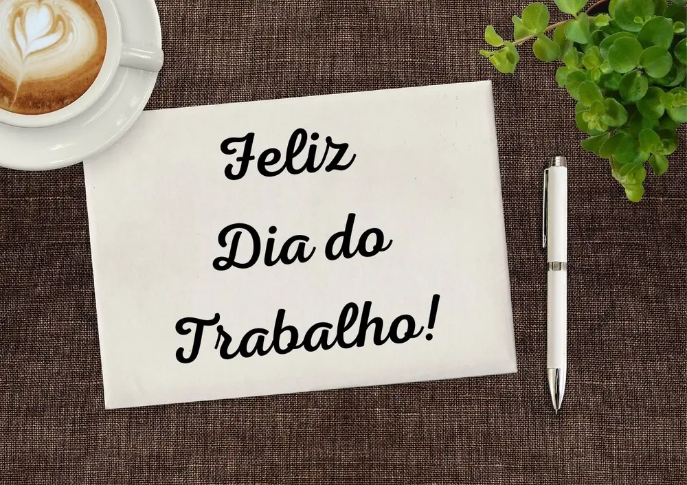 Foto de mesa com xícara de café, planta, caneta e folha escrito "feliz dia do trabalho"