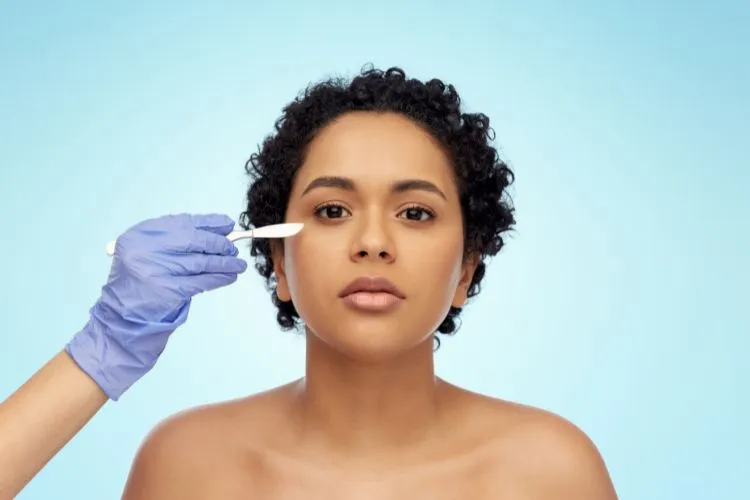 Mulher de pele negra e cabelo cacheado curto, com mão de outra pessoa usando luvas azuis e segurando bisturi próximo ao olho dela