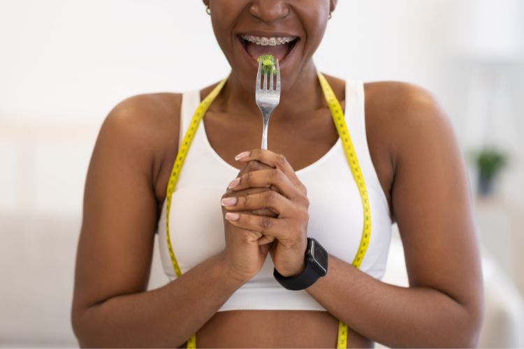 A imagem mostra uma pessoa segurando um garfo com um pedaço de comida verde, possivelmente um vegetal. A pessoa está usando uma fita métrica amarela ao redor do pescoço, sugerindo um tema de dieta ou saúde. Além disso, a pessoa está usando um relógio preto no pulso esquerdo. A pessoa está vestindo uma blusa branca sem mangas e o fundo da imagem é claro e minimalista, colocando o foco na pessoa e no garfo com comida.