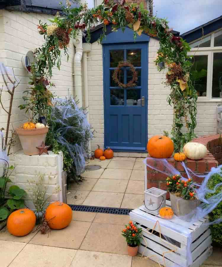 Entrada de casa com tijolos à vista, porta azul com guirlanda seca, plantas com teias de aranha e abóboras 