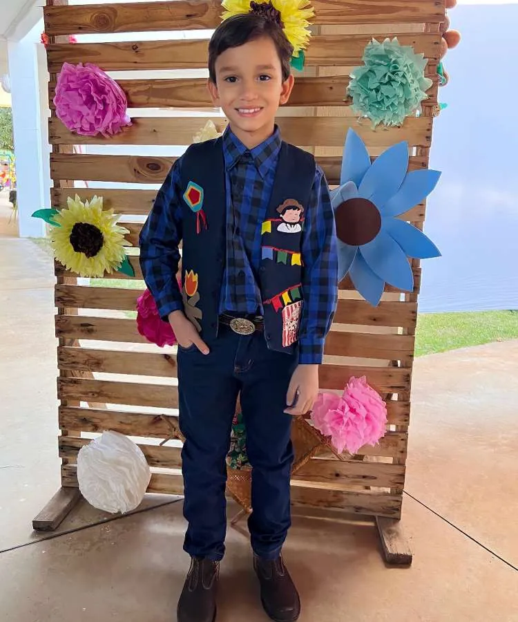 Menino usando camisa xadrez azul, colete, calça jeans e sapatão para festa junina.
