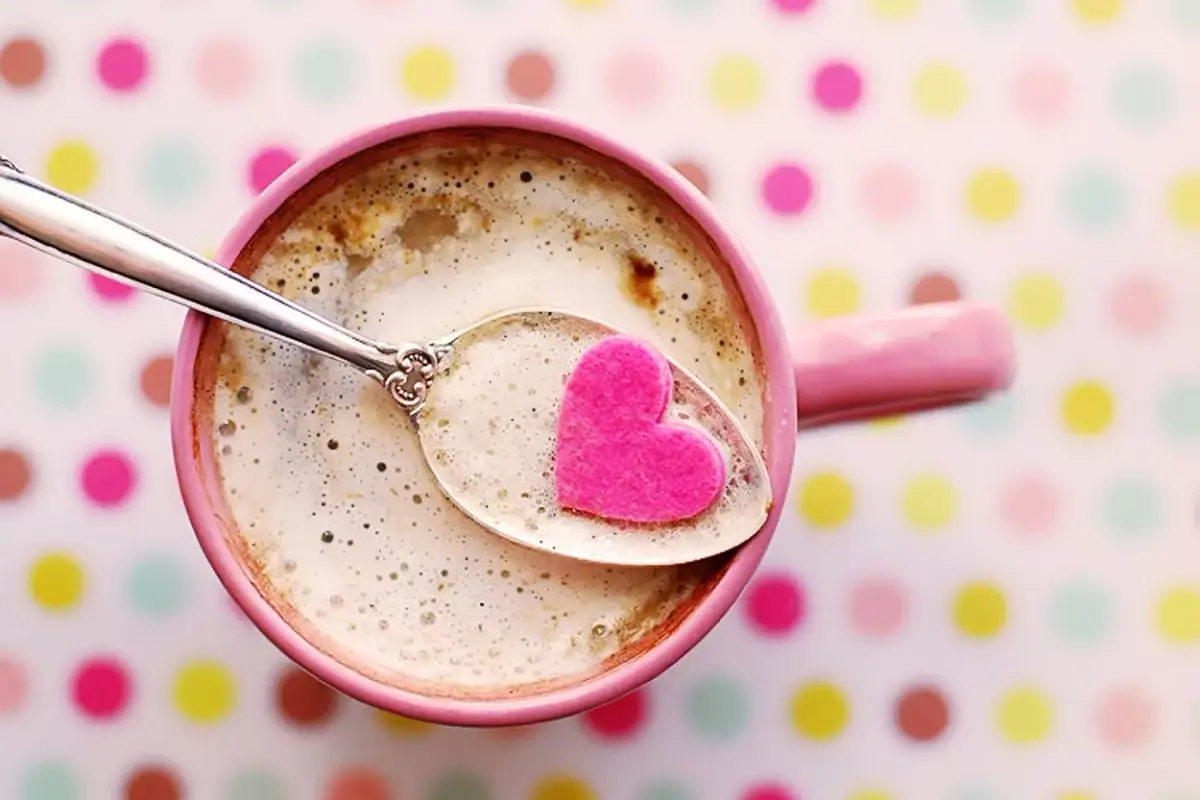 Foto de xícara rosa de cafe, colher prata com café e coração rosa. Ao fundo, a mesa é branca de bolinhas coloridas 