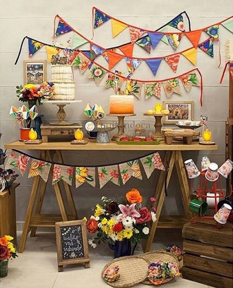 A imagem representa decoração para festa junina com mesas diferentes. Contém doces, bandeirinhas, arranjos de flores, peneiras depalha, plaquinhas, etc