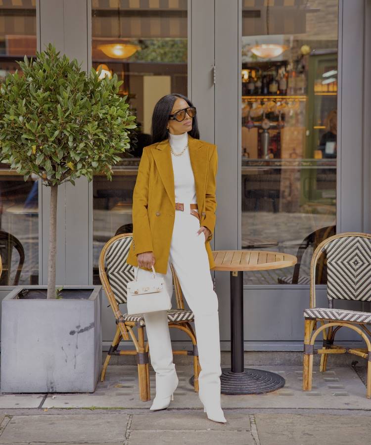 Mulher estilosa em frente a um café, veste blazer mostarda, blusa branca de gola alta, calça branca e botas combinando. Usa cinto marrom, óculos de sol grandes e segura bolsa branca. Mesas, cadeiras e planta no ambiente externo.