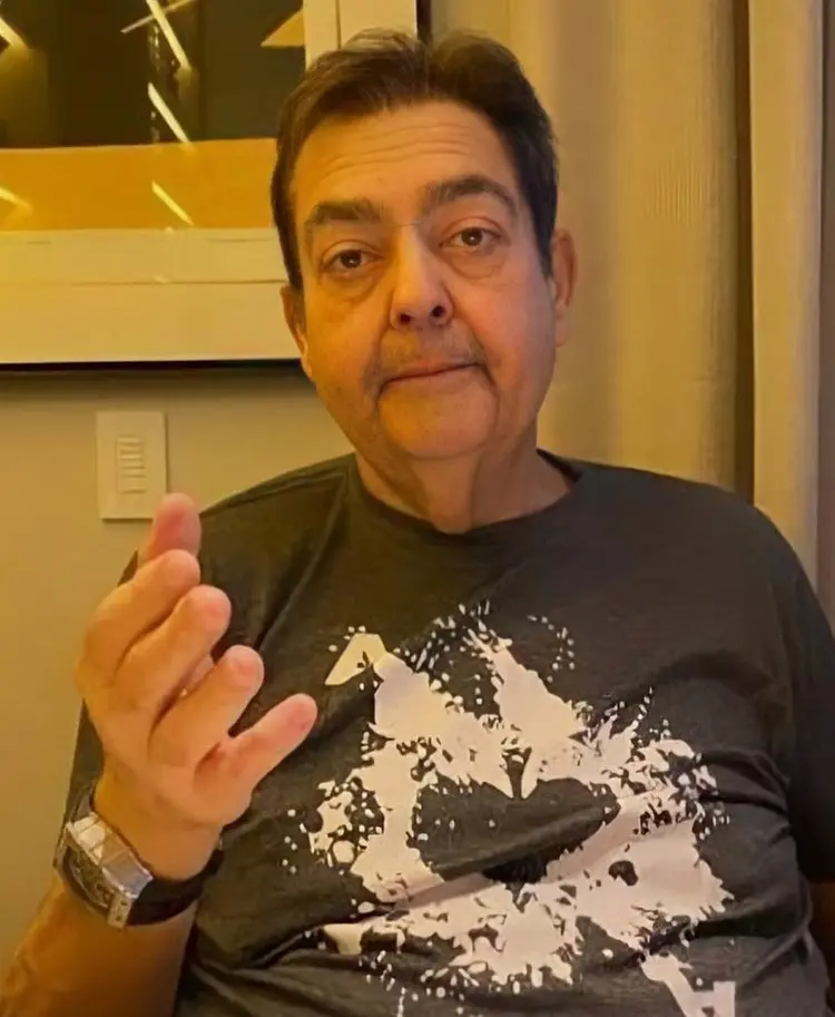 Apresentador Fausto Silva, usando camiseta cinza, com a mão entreaberta, sentado em frente à parede com quadro, itnerruptor e cortina
