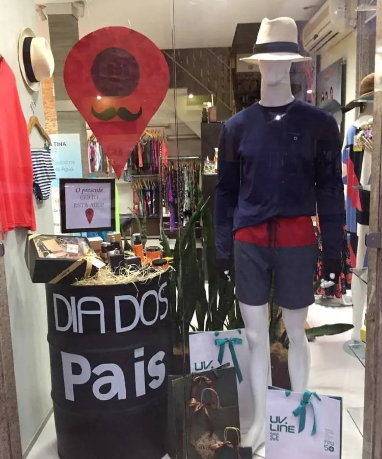 A imagem mostra uma vitrine de loja com foco no Dia dos Pais. Um manequim veste roupa de verão, e há um barril decorado com "DIA DOS Pais" exibindo produtos. Um balão vermelho em formato de pino com bigode e um chapéu adornam a vitrine.