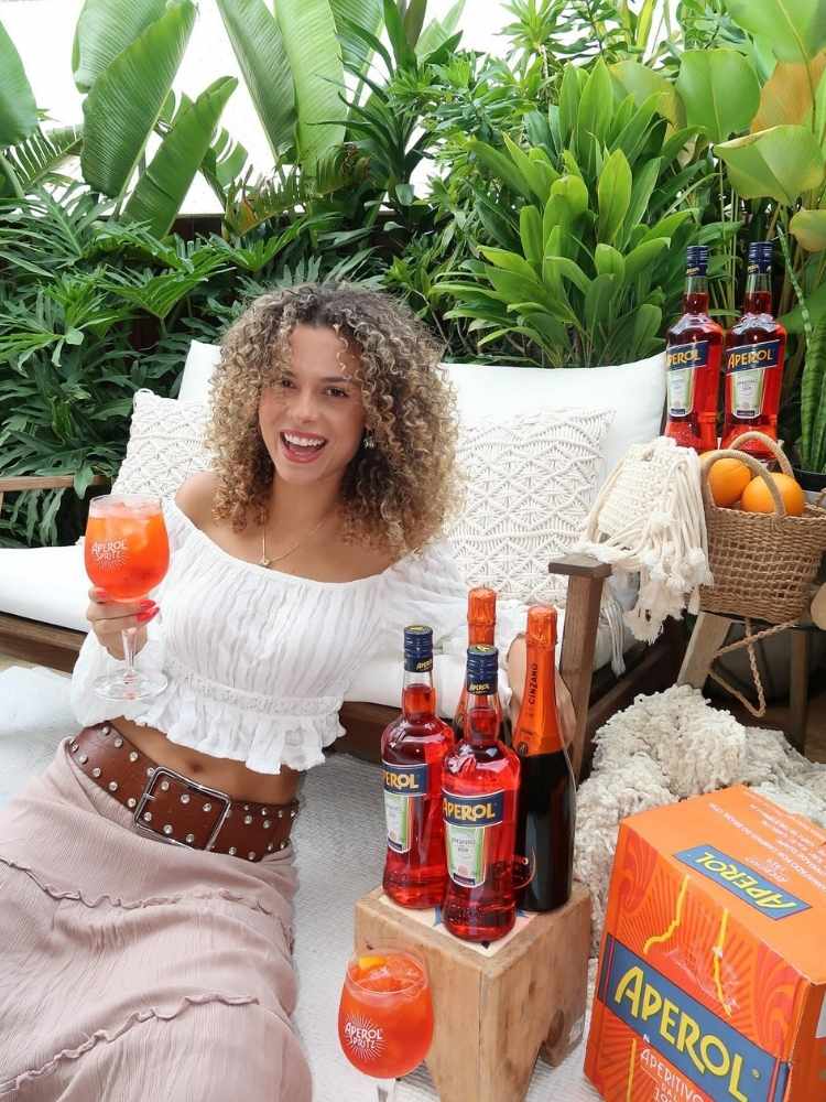 Larissa usando cropped branco, saia bege com cinto marrom, sentada em sofá com garrafas e fardos de Aperol