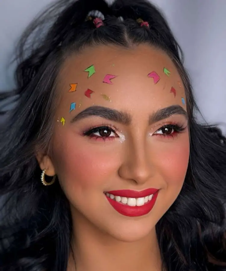 Uma mulher sorridente com maquiagem de Festa Junina, destacando olhos com sombras em tons de rosa e branco. Pequenas bandeirinhas coloridas e confetes adornam sua testa, e seus lábios estão pintados com um batom vermelho vibrante.