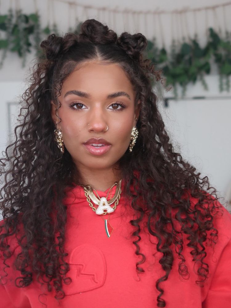 Mulher de pele negra com cabelo castanho cacheado com 3 coques no topo e resto solto 