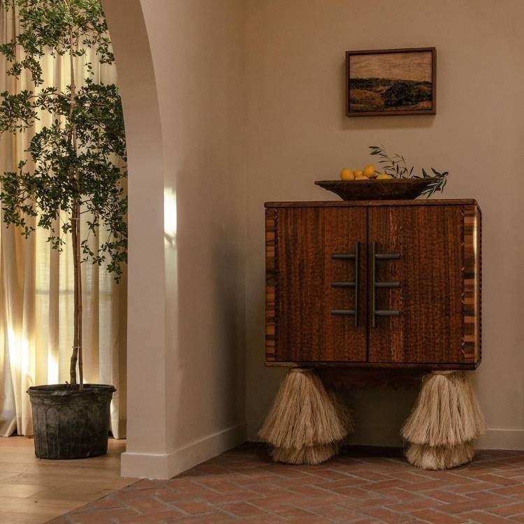 A imagem apresenta um ambiente minimalista e rústico. Um armário de madeira com detalhes únicos em palha nas bases destaca-se, decorado com uma tigela de madeira contendo limões e ramos verdes. Ao lado, uma planta em vaso metálico complementa a estética natural.