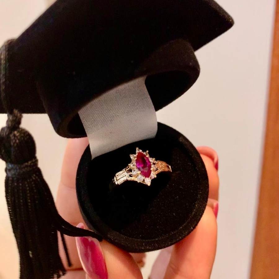 Pessoa de pele clara segurando caixa de joia em formato de chapéu de formatura, com anel de rubi dentro
