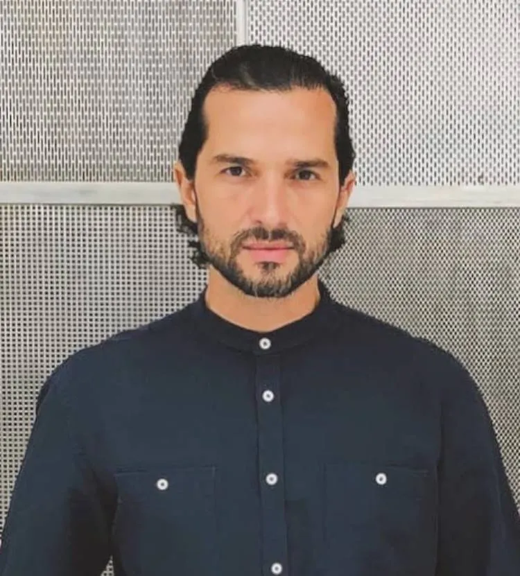 Ator Jeff Machado, com camisa social azul-escuro de frente a placa de metal com furinhos
