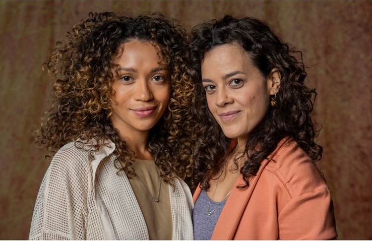 Laís e Cecília de Vale Tudo com cabelos cacheados estão lado a lado. A mulher da esquerda tem cachos mais claros e usa uma camisa clara sobre uma blusa neutra. A mulher da direita tem cachos escuros, usa um blazer cor de pêssego sobre uma blusa lilás, e brincos. Ambas olham para frente com um leve sorriso.
