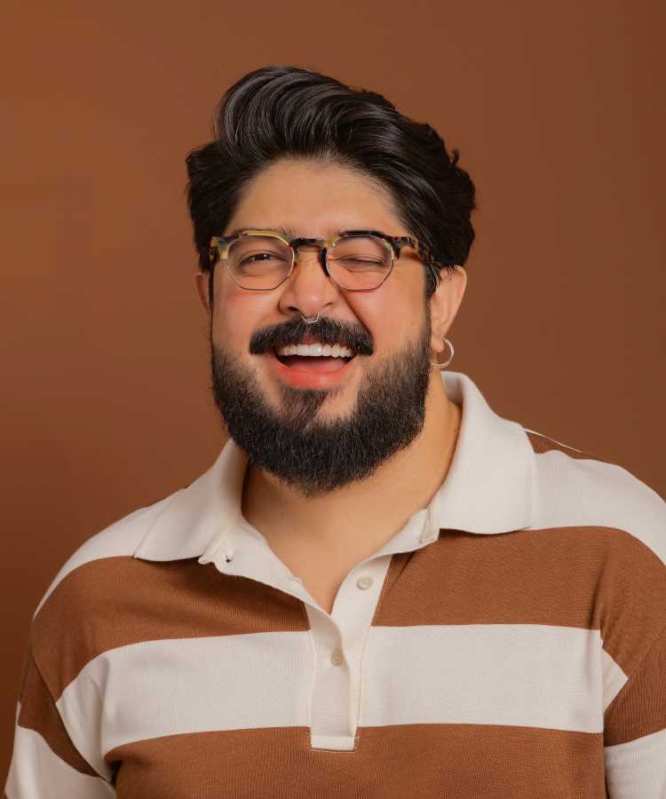 Yan Yoshimitsu Sato orridente com barba cheia e óculos de armação estampada posa contra fundo marrom. Ele usa camisa polo listrada em branco e marrom, transmitindo um estilo casual e descontraído em um retrato iluminado com cores quentes.