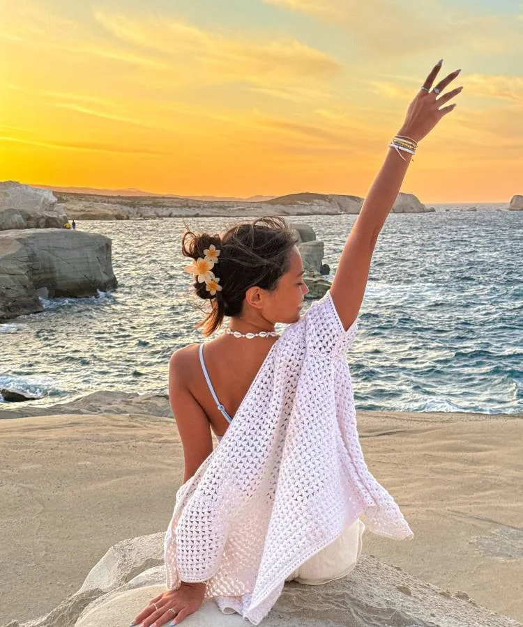 Uma mulher de cabelos presos com flores brancas, usando um top azul e um casaco de crochê branco, está sentada em uma rocha à beira-mar. Ela levanta o braço em direção ao céu alaranjado do pôr do sol. Fotos tumblr na praia capturam momentos assim.


