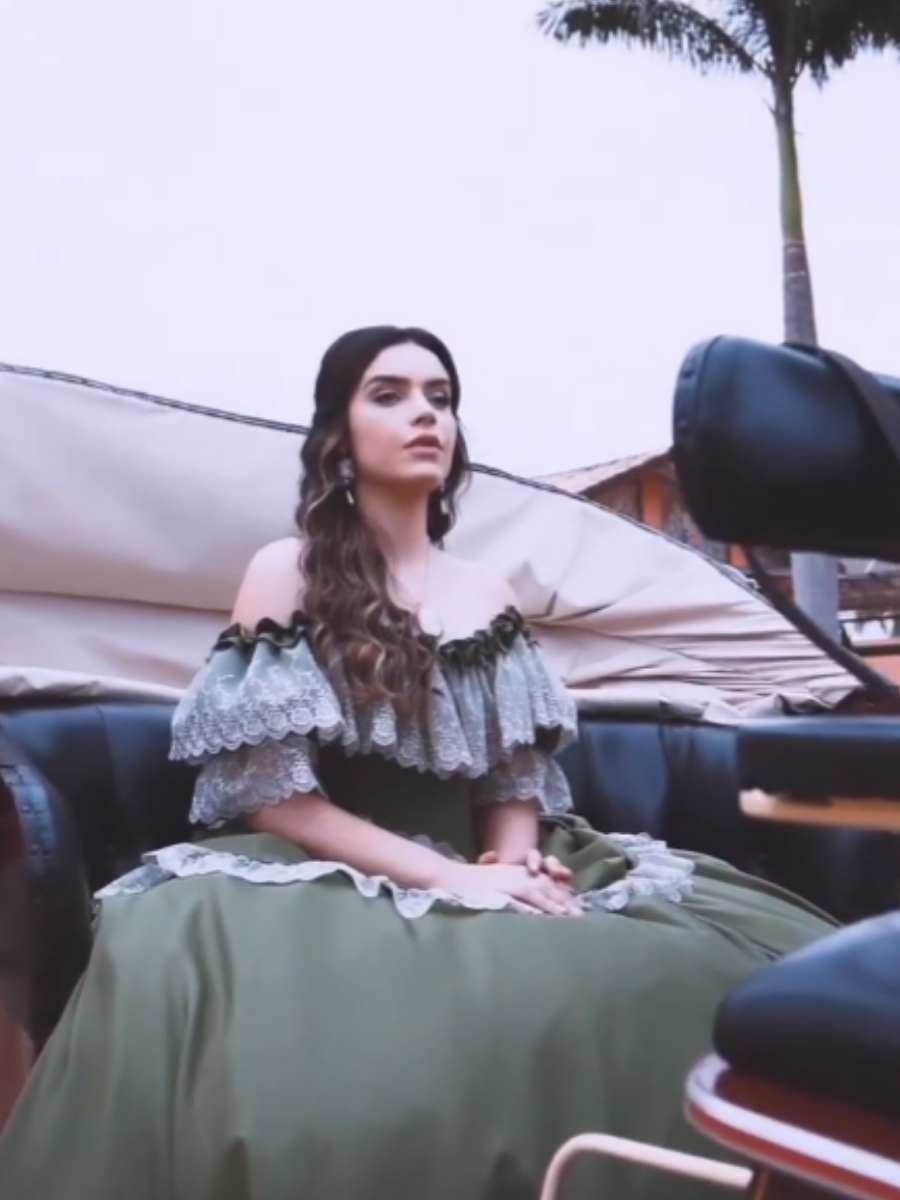 Imagem em fundo com céu, palmeira e carruagem. Karol está sentada na carruagem fantasiada de Katherine da série "The Vampire Diaries" para o Halloween Sephora. Ela veste vestido verde com babados em renda. 