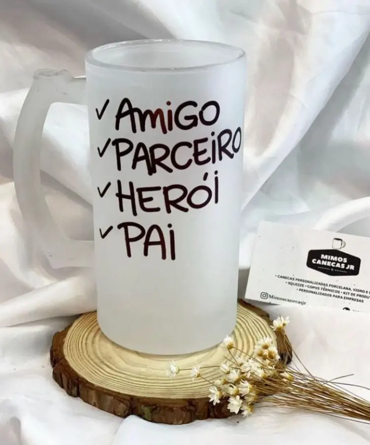 Caneca branca com palavras “Amigo, Parceiro, Herói, Pai” escritas verticalmente. Apresentada sobre um corte de tronco de árvore e decorada com pequenas flores brancas e talos secos.