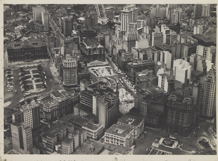 A imagem aérea retrata o centro de São Paulo em meados do século XX, com prédios históricos, ruas movimentadas e a Catedral da Sé ao fundo. A urbanização crescente contrasta com espaços abertos, destacando o dinamismo urbano da época.