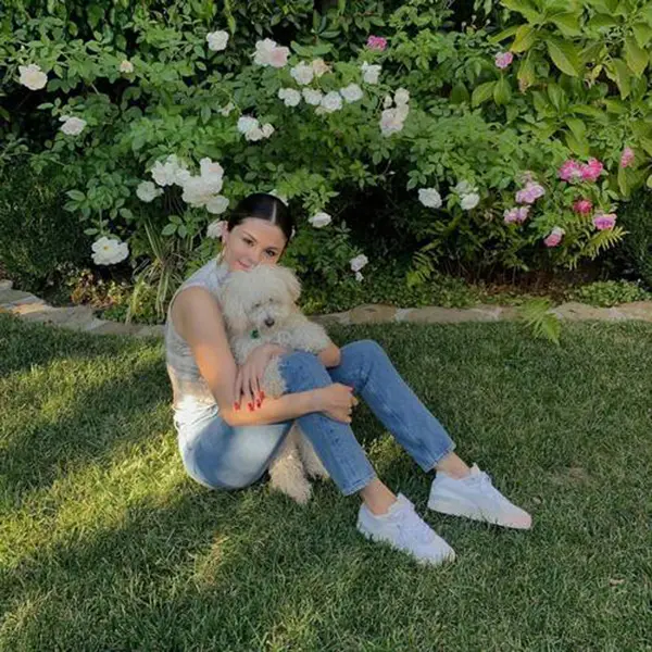 Selena Gomez de regata cinza, calça jeans e tênis branco sentada na grama com poodle branco