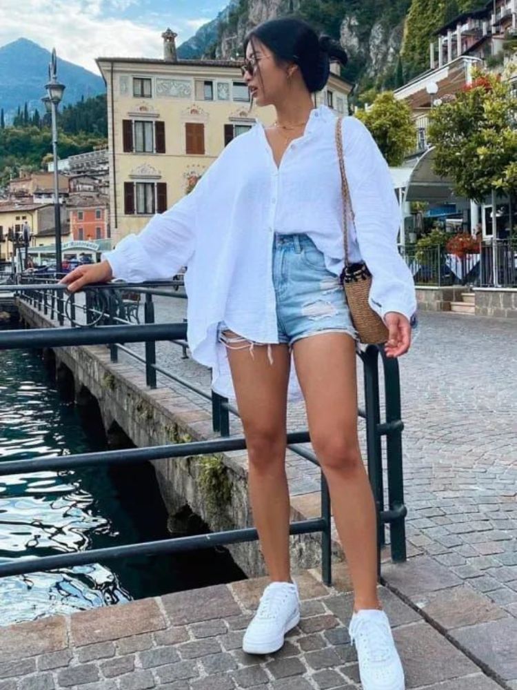 Mulher de pele clara com look com camisa social branca, shorts jeans e tênis branco.