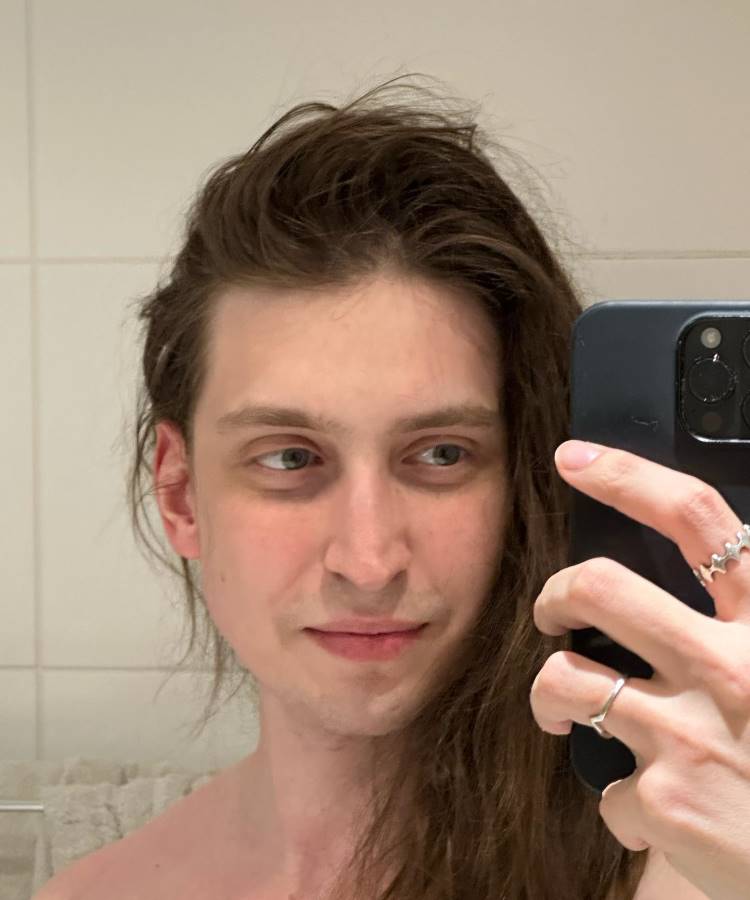 Felca de cabelo castanho comprido e bagunçado, com o tronco nu, tirando uma selfie no espelho do banheiro. Ele segura um celular com uma mão que exibe anéis. Ele olha para a câmera com um sorriso discreto.