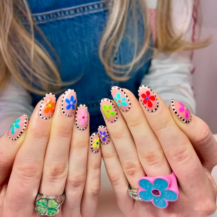 A imagem mostra um close-up de duas mãos com os dedos estendidos, exibindo arte detalhada nas unhas. Cada unha apresenta um design único com uma variedade de cores e padrões. Os designs incluem motivos florais com pétalas e centros de cores diferentes, bolinhas e contornos acentuados ao redor das unhas. As cores são vibrantes e incluem tons de rosa, laranja, azul, verde, amarelo e preto. Algumas unhas têm uma única flor grande, enquanto outras têm várias flores menores ou pontos.O fundo está desfocado, mas parece ser um ambiente interno com tons rosados. A pessoa cujas mãos são mostradas está vestindo uma peça de roupa jeans, sugerindo um traje casual.