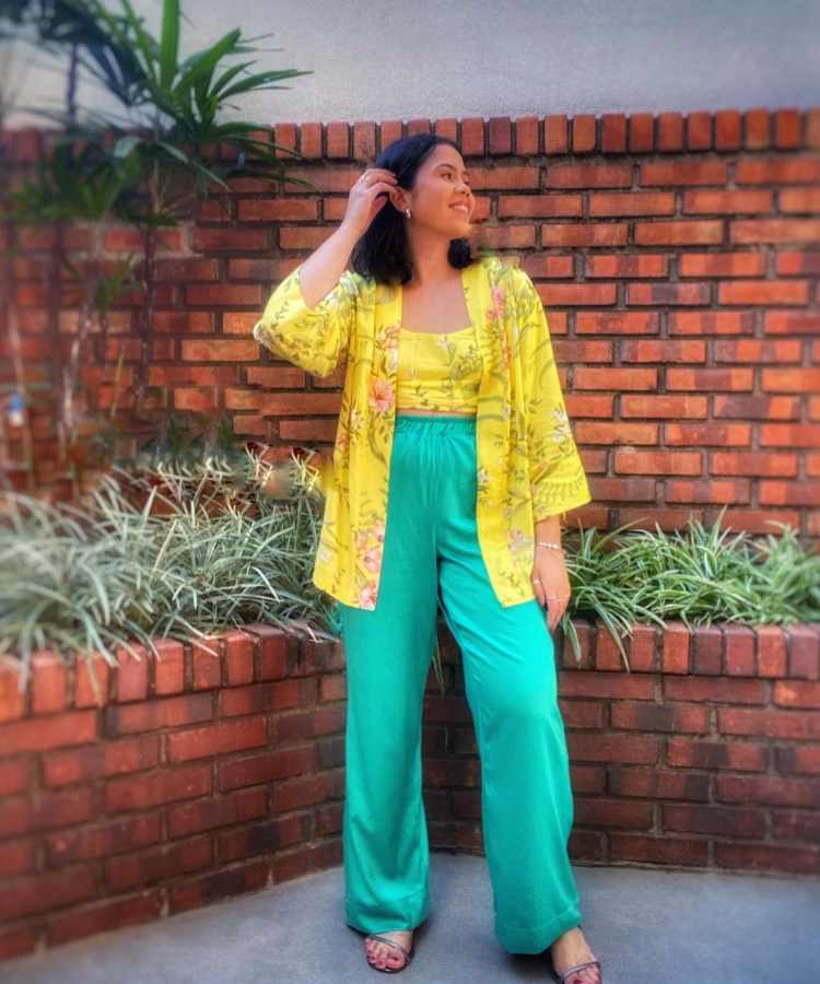 Imagem com fundo de parede de tijolinhos. No centro, mulher usando camisa + cropped amarelo estampado + calça verde