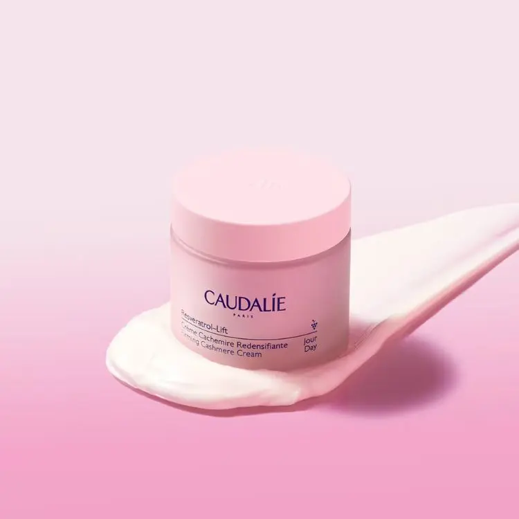 A imagem apresenta um pote rosa pastel de Caudalie Resveratrol-Lift Crème Cachemire Redensifiante, um creme diurno. O pote está sobre uma camada branca de produto, que se estende para a direita, em um fundo rosa que se degrada para um tom mais claro.

