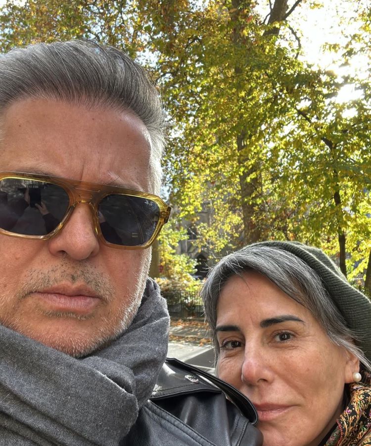 Gloria Pires e Orlando Morais, Um casal maduro posa para uma selfie ao ar livre em um dia ensolarado de outono. O homem usa óculos escuros, jaqueta de couro e cachecol; a mulher, de gorro verde e casaco, sorri suavemente.