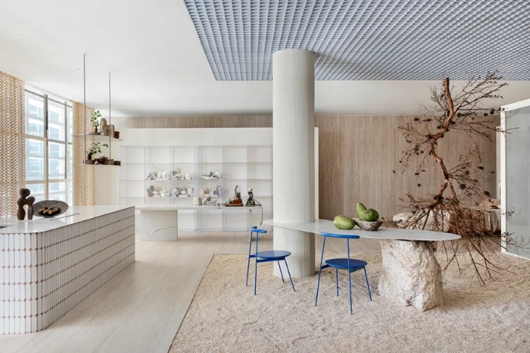 A imagem mostra uma sala de estar com estilo moderno, paredes de tijolos aparentes laranjas, sofá bege, poltronas marrons e brancas, mesa de centro geométrica em madeira e decoração minimalista.

