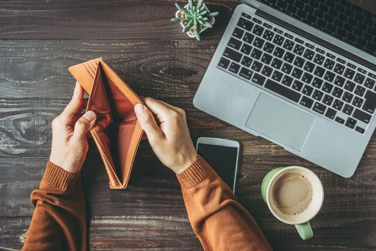 Uma pessoa segura uma carteira vazia sobre uma mesa de madeira, ao lado de um laptop, um celular e uma xícara de café. A imagem transmite a sensação de que é preciso buscar formas de sair do vermelho financeiramente.

