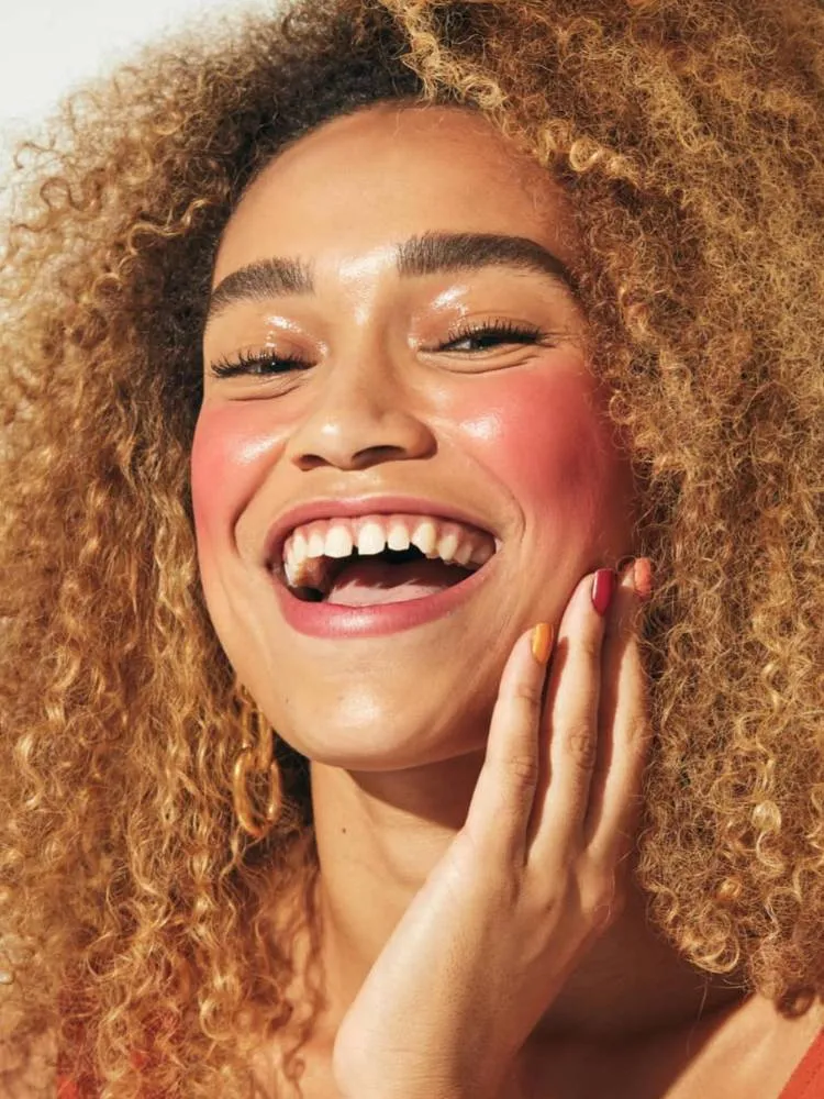 Mulher de pele clara e cabelo cacheado usando bastante blush e gloss transparente nas pálpebras