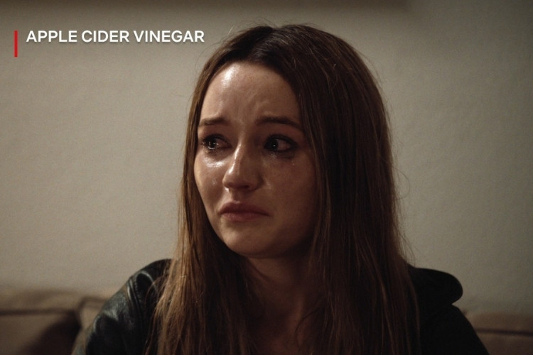 Kaitlyn Dever, interpretando Belle Gibson na série "Vinagre de Maçã" da Netflix, aparece emocionada, com lágrimas escorrendo pelo rosto. Ela está em um ambiente interno, com expressão de tristeza, usando uma jaqueta escura. O título da série está visível.


