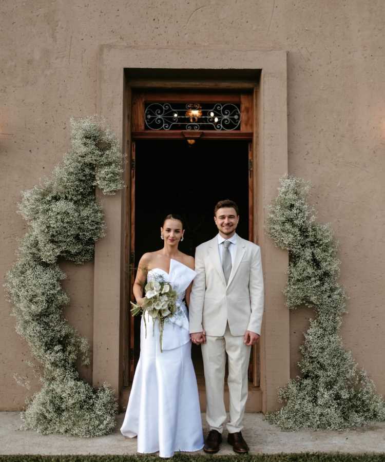 Casal vestido para casamento posando em frente a porta de madeira ornamentada com folhagens delicadas. A noiva em vestido branco com bouquet, e o noivo de terno claro, transmitem elegância e simplicidade em ambiente rústico e romântico.