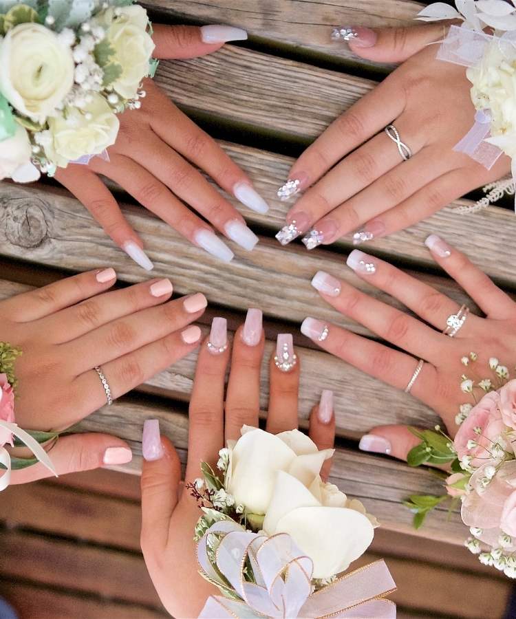 5 mãos de mulheres de pele clara sobre mesa de madeira, com unhas quadradas de diversos tamanhos, envolvendo técnicas como glitter, pedrarias e esmalte fosco