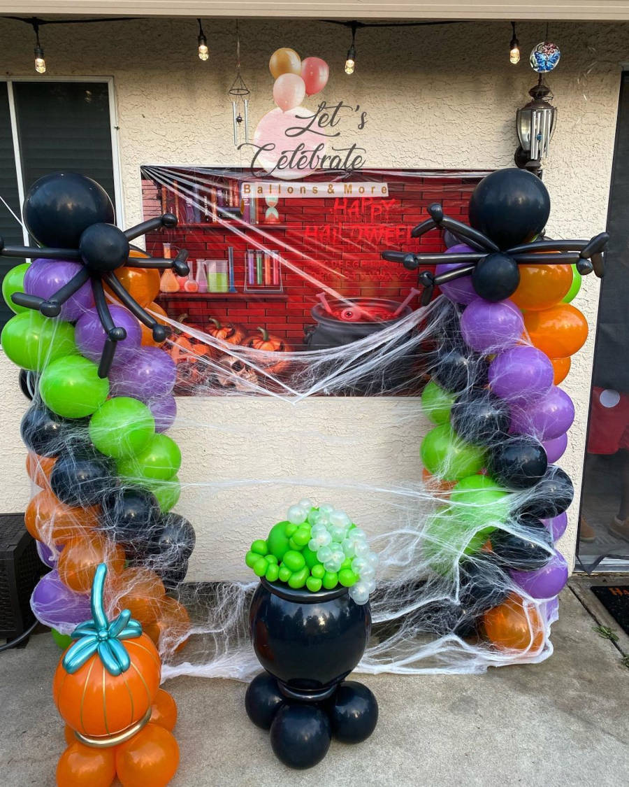 Decoração com bexigas em preto, verde, roxo e laranja, painel em vermelho com desenho de caldeirão escrito "happy halloween" e teias de algodão.