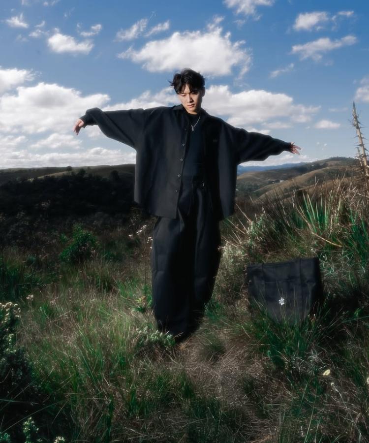 Modelo de pé com braços abertos usando look all black: camisa oversized, camiseta e calça baggy