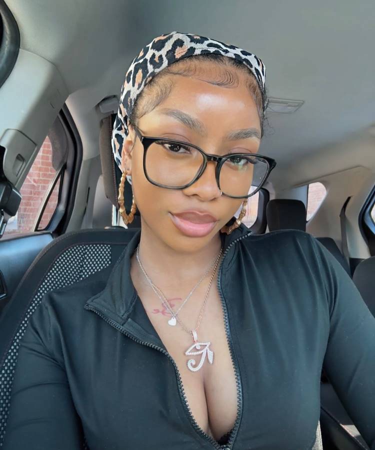Mulher dentro de um carro, usando óculos grandes, brincos de argola dourados e lenço com estampa animal print. Baby hairs estilizados na testa, maquiagem natural, colar brilhante e zíper da blusa preta parcialmente aberto, com expressão confiante.