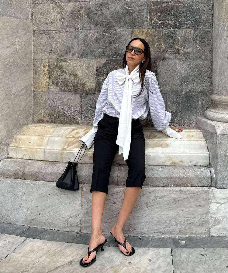 Uma mulher com uma elegante blusa de listras finas, um grande laço branco no pescoço e calças pretas capri. Ela está sentada em um banco de pedra, usando chinelos de salto preto e carregando uma bolsa pequena.