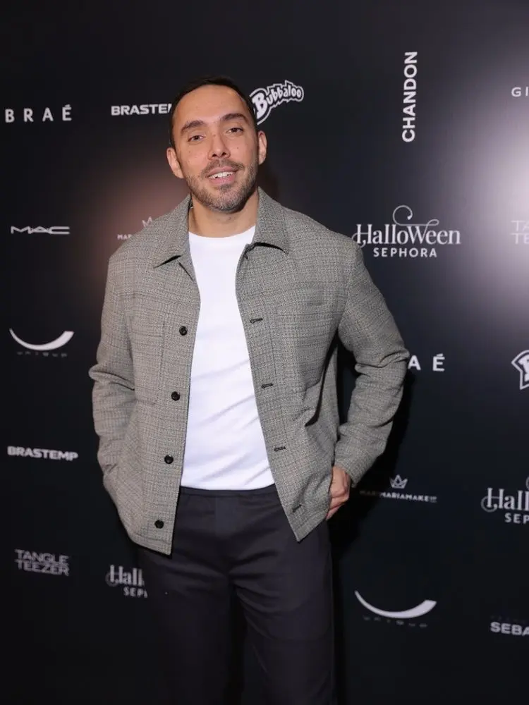 A imagem mostra um homem sorrindo em frente a um painel de marcas. Ele tem cabelo curto e barba. Veste uma jaqueta cinza sobre uma camiseta branca e calças pretas. O fundo é preto com logotipos de várias marcas em branco, incluindo "Halloween da Sephora 2024".


