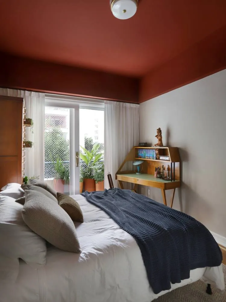 Esta imagem mostra um quarto com uma cama de casal e roupa de cama branca, coberta por um cobertor azul-marinho. O teto é pintado de terracota, contrastando com as paredes claras. Uma escrivaninha de madeira está ao lado da cama, e uma porta de vidro se abre para uma varanda com plantas.

