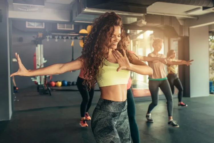 Mulher de cabelo ondulado e roupa fitness ao lado de outras mulheres dançando em academia com equipamentos de exercício no fundo